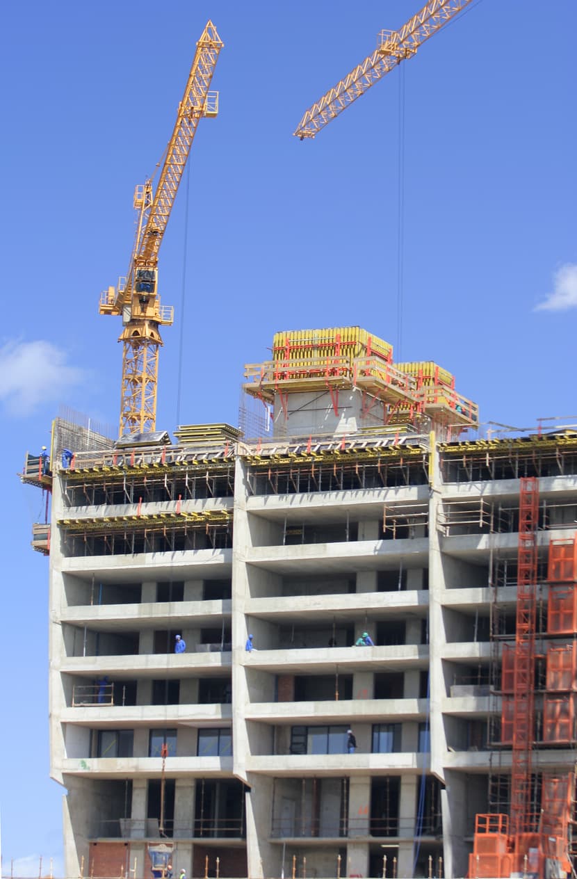 Construcción de un edificio en proceso, con grúas y trabajadores en diferentes niveles de la estructura de concreto. El cielo está despejado.