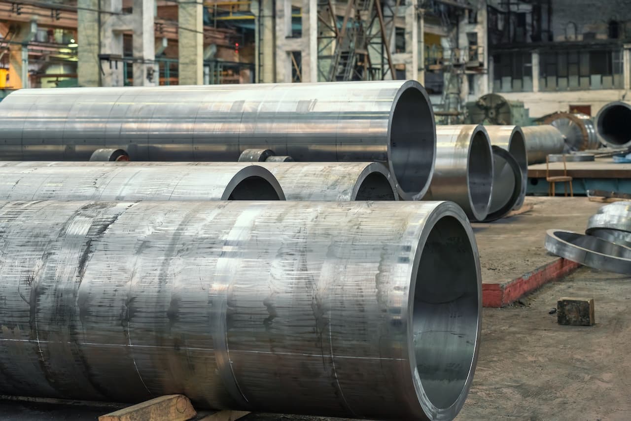 tubería de acero inoxidable apilada en una industria, con un entorno de trabajo visible y estructura metálica de fondo.